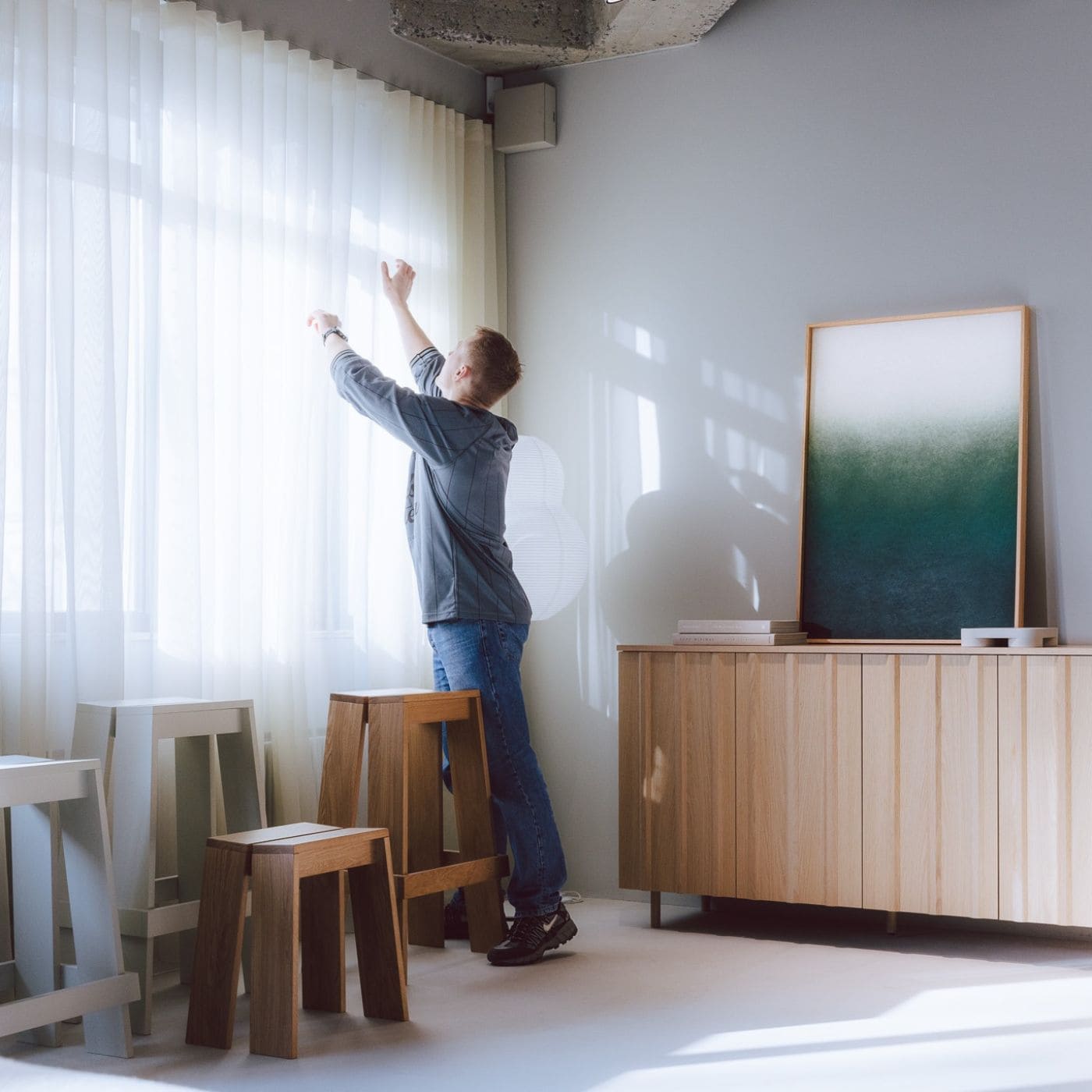 Mand justerer gardiner i et lyst rum hvor et akustisk billede med grønne nuancer skaber ro og forbedrer akustikken i indretningen.