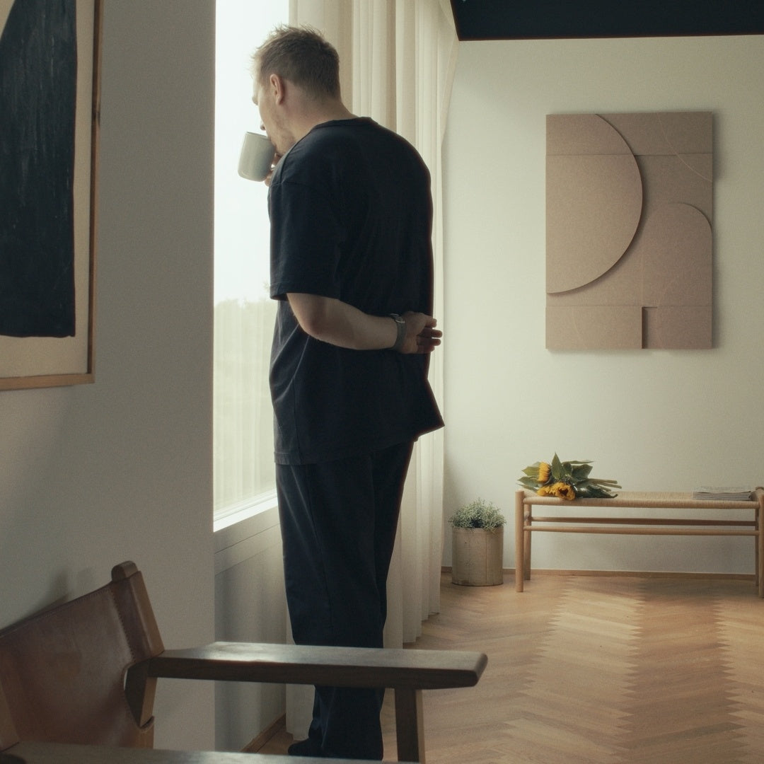 Mand, der står ved et vindue i et roligt, minimalt rum med akustisk vægkunst designet til bedre rumakustik.